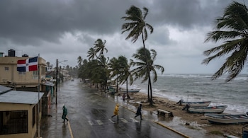 Calle costera inundada con personas en impermeables. Palmeras se agitan, mar picado y botes en la orilla bajo un cielo oscuro y una bandera dominicana.