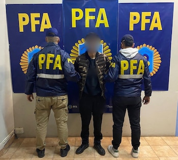 Dos oficiales de la PFA, vistos de espaldas con chaquetas azules que dicen "PFA", escoltan a una persona con una chaqueta de camuflaje, frente a tres estandartes azules con el logo de la PFA