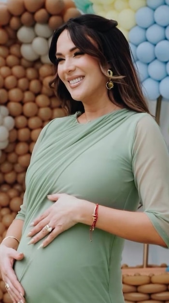 Una mujer sonriente con vestido verde apoya sus manos en su vientre gestante ante un fondo decorado con globos
