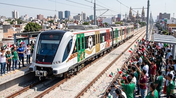 Un tren blanco y verde decorado con balones de fútbol avanza sobre una vía elevada. Abajo y a los lados, una multitud de personas ondea banderas mexicanas.