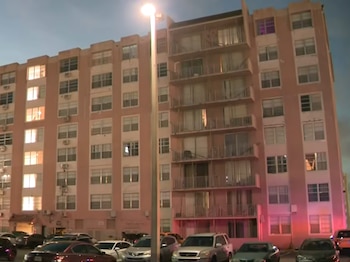 Edificio residencial rosa claro con ventanas iluminadas y balcones, visto de noche. Un poste de luz y varios coches, incluyendo SUVs, están estacionados