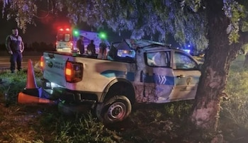 Una camioneta oficial dañada junto a un árbol al costado de una ruta de noche, con conos de tránsito y una ambulancia al fondo