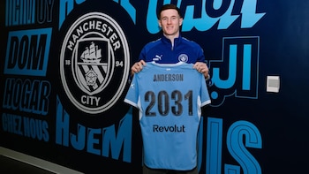Hombre sonriente con chaqueta azul sostiene camiseta celeste de fútbol con "ANDERSON" y "2031" sobre fondo oscuro con logo del Manchester City y textos