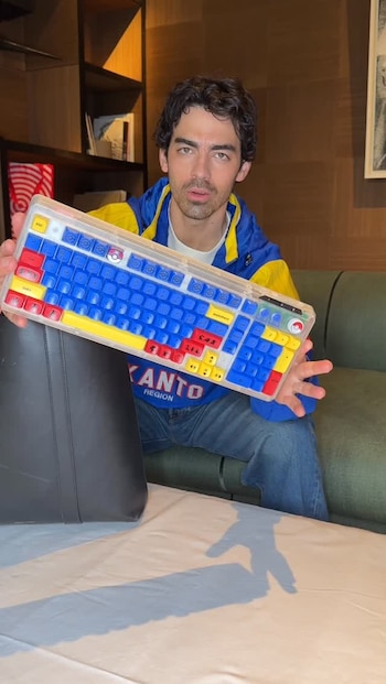 Hombre con chaqueta azul y amarilla que dice 'KANTO REGION' sostiene un teclado transparente de colores. Se ve estantería y sofá verde en una habitación