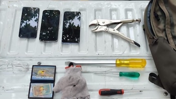 Tres teléfonos, pinzas de presión, tres destornilladores, gafas de seguridad, una cartera, un guante y parte de una mochila sobre una superficie blanca