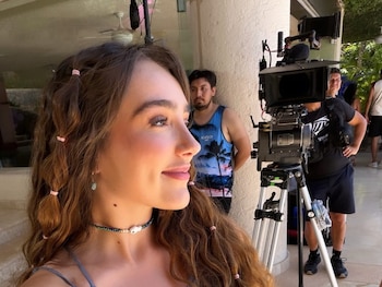 Una mujer joven con cabello largo y ondulado, top de encaje azul y falda floral, sonríe en un primer plano. Equipo de filmación y personal al fondo