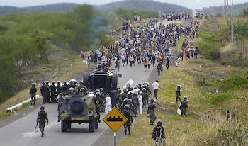Vista aérea de una carretera con una gran multitud de personas, soldados con equipo antidisturbios y vehículos militares, con humo visible y colinas al fondo