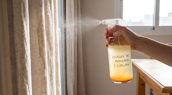 Mano que sostiene un atomizador transparente con líquido de vinagre de manzana y cúrcuma, rociando una cortina de tela beige.