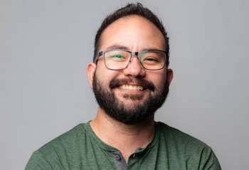Hombre sonriente con barba, gafas y camiseta verde sobre un fondo gris neutro