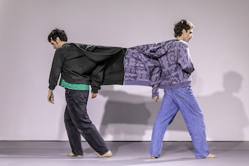 Dos hombres descalzos unidos por chaquetas con cremallera, uno de negro y otro de morado estampado, en una sala con paredes claras