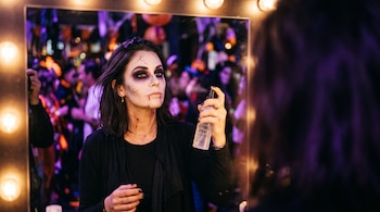 Reflejo en un espejo de una mujer maquillada con sombras oscuras y detalles faciales, que sostiene un frasco atomizador frente a su rostro.