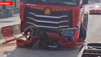Primer plano de la cabina de un camión rojo con el parachoques y la parrilla destrozados, junto a barreras de tráfico en una carretera. Un coche rojo al fondo
