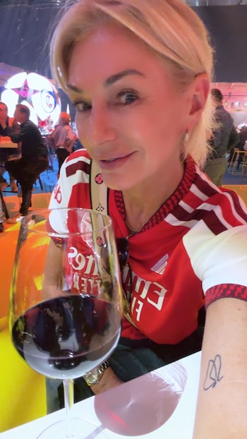Yanina Latorre, una mujer rubia, posa con una camiseta roja y blanca del Arsenal y una copa de vino tinto. Se ve un tatuaje en su muñeca