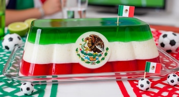 Gelatina tricolor (verde, blanco, rojo) con el escudo de México en el centro, decorada con pequeñas banderas mexicanas y balones de fútbol.
