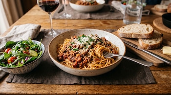 Plato de fideos con salsa de carne y queso, ensalada verde, rebanadas de pan y una copa de vino tinto sobre una mesa de madera.
