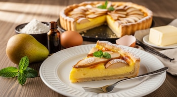 Primer plano de una porción de tarta de peras con crema pastelera y menta. Al fondo, la tarta entera, una pera, harina, huevo, extracto de vainilla y mantequilla.
