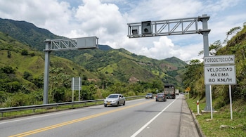 Vista de una carretera colombiana con varias cámaras de fotodetección sobre un pórtico metálico, vehículos circulando y montañas verdes al fondo bajo un cielo nublado.
