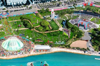 Vista aérea conceptual de un festival masivo en Bayfront Park junto al agua, con gente, pantallas gigantes, escenarios, estructuras de patrocinadores y zonas verdes