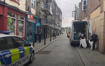 Vista de una calle urbana con un coche de policía a la izquierda, dos agentes de policía, y una furgoneta blanca con personal manipulando bolsas