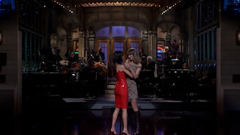 Dos mujeres sobre un escenario iluminado, una con vestido rojo y otra con vestido a cuadros, frente a una banda de música al fondo