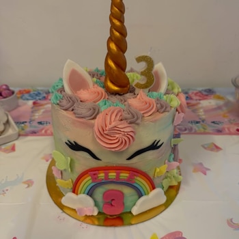 Torta de cumpleaños con forma de unicornio, con un cuerno dorado, orejas, ojos cerrados, glaseado de colores pastel, mariposas, un arcoíris, el nombre ELLE y un número 3