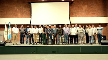 Grupo de diecinueve personas de pie sobre un escenario, con un telón de fondo de ladrillo, una pantalla blanca, dos banderas y el logotipo de ANCAFE