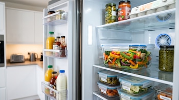 Interior de un refrigerador abierto con recipientes de vidrio que contienen verduras, frascos de conservas, botellas y un cartón de huevos.