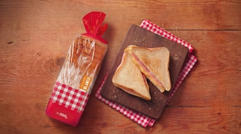 Vista aérea de un paquete de pan blanco y un sándwich de jamón y queso cortado por la mitad sobre una tabla de madera y un mantel a cuadros rojo y blanco.