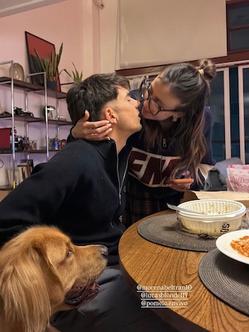 Una mujer con gafas se inclina hacia un hombre sentado a una mesa de comedor para darle un beso, con un perro dorado en primer plano