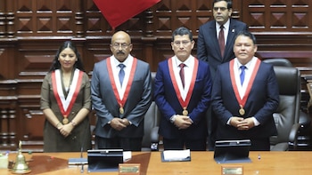 Cuatro personas, tres con bandas bicolores rojas y blancas y medallas, de pie en un salón con paneles de madera. En primer plano hay una mesa con objetos