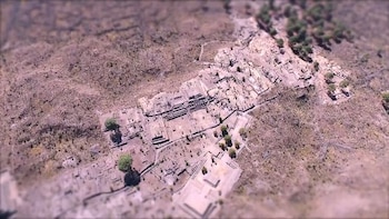 Vista aérea de un complejo arqueológico con ruinas de piedra, árboles pequeños y vegetación dispersa en un terreno seco y rojizo