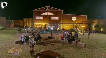 Escena nocturna de La Granja VIP con concursantes sentados en un patio de césped frente a una casa de madera iluminada, con un estanque y un cartel del programa