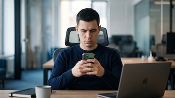Hombre en una oficina sentado en su silla, usa un teléfono móvil, con un ordenador portátil, una libreta y una taza de café en el escritorio