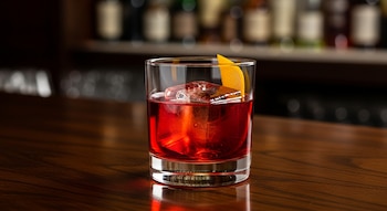 Vaso de cristal con cóctel Negroni rojo, un cubo de hielo grande, una cáscara de naranja y fondo borroso con botellas en una barra de madera oscura.