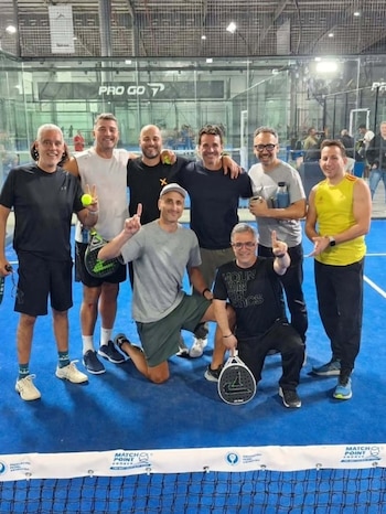Ocho hombres con indumentaria deportiva posan dentro de una cancha de pádel de suelo azul, frente a una red y paneles de vidrio