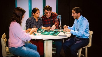 Cuatro personas sentadas en una mesa redonda juegan Scrabble con tablero, fichas, atriles, vasos con agua y un teléfono celular; un andador detrás de un jugador