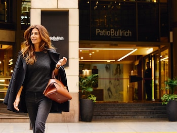 Mujer de cabello castaño largo caminando por una acera. Viste un abrigo oscuro y jeans, llevando un bolso marrón. Al fondo se ven tiendas de lujo