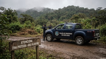 Una camioneta de la Policía de Fronteras de Costa Rica en camino de tierra, dos oficiales con binoculares. Vegetación densa, montañas neblinosas y letrero de zona protegida.