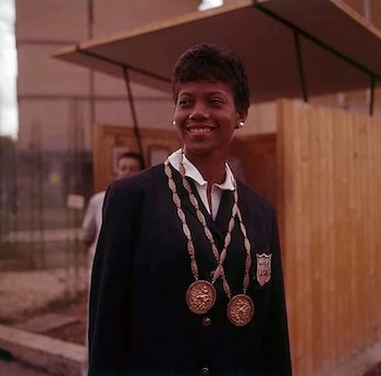 Retrato de una mujer afroamericana sonriendo, con chaqueta oscura, camisa blanca, dos medallas grandes colgando y un escudo de Estados Unidos