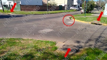 Intersección con asfalto agrietado y parches. Se ven aceras, césped y rejillas de desagüe pluvial. Flechas rojas y un círculo señalan el deterioro visible