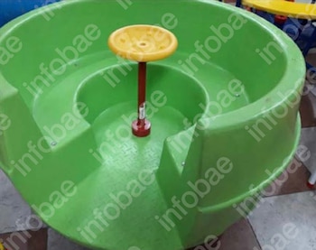 Calesita de juego infantil de plástico, color verde. Posee un poste central rojo que sostiene un volante o asiento circular amarillo. Fondo con baldosas