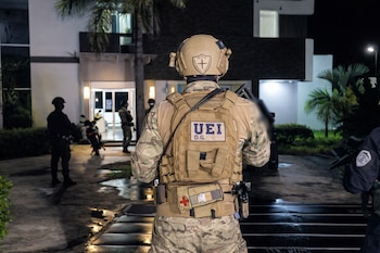 Vista trasera de un agente con uniforme de camuflaje y chaleco táctico "UEI D.G.", parado frente a un edificio moderno de noche. Otros agentes y una motocicleta son visibles.