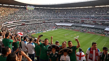 Estadio de fútbol con gradas llenas de aficionados. Un marcador electrónico muestra el partido México contra Inglaterra. En primer plano, aficionados celebran con banderas y sombreros.