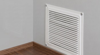 Una rejilla de ventilación blanca con lamas horizontales y cuatro tornillos está en una pared gris junto a un zócalo y un piso de madera.