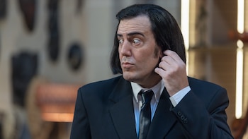 Un hombre de mediana edad con cabello oscuro liso y patillas, vestido con traje y corbata negros, con expresión sorprendida y mano en la oreja