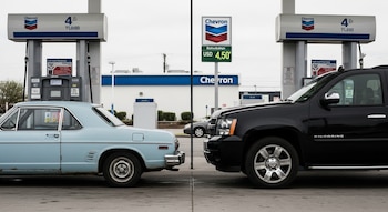 Un auto azul claro antiguo y un SUV negro moderno en surtidores separados de una estación Chevron con un cartel que muestra el precio de 4.50 USD por galón.