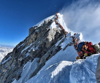 Una larga fila de alpinistas con ropa de colores brillantes y mochilas asciende por una empinada arista nevada y rocosa del Monte Everest bajo un cielo azul claro