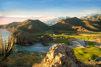 Vista aérea de montañas cubiertas de vegetación, océano, campo de golf con lagunas y puentes, y cactus en primer plano, bajo cielo azul