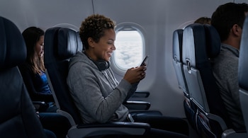 Mujer sonriente de pelo rizado y tez oscura con sudadera gris y auriculares, mirando su celular en un asiento de avión junto a la ventana que muestra nubes.