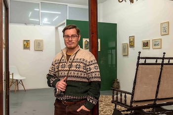 Hombre con gafas y jersey estampado sostiene una cámara fotográfica en una sala con obras de arte enmarcadas y mobiliario antiguo